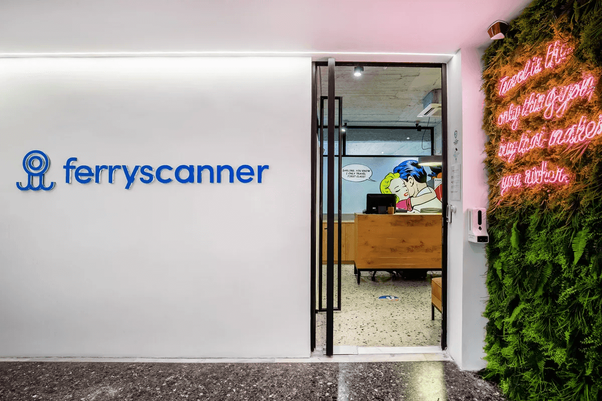 The Ferryscanner office entrance featuring a white wall with the Ferryscanner logo, a glass door leading into the lobby with pop art wall art, and a green vertical garden wall to the right illuminated by a pink neon sign that reads, "Travel is the only thing you buy that makes you richer".