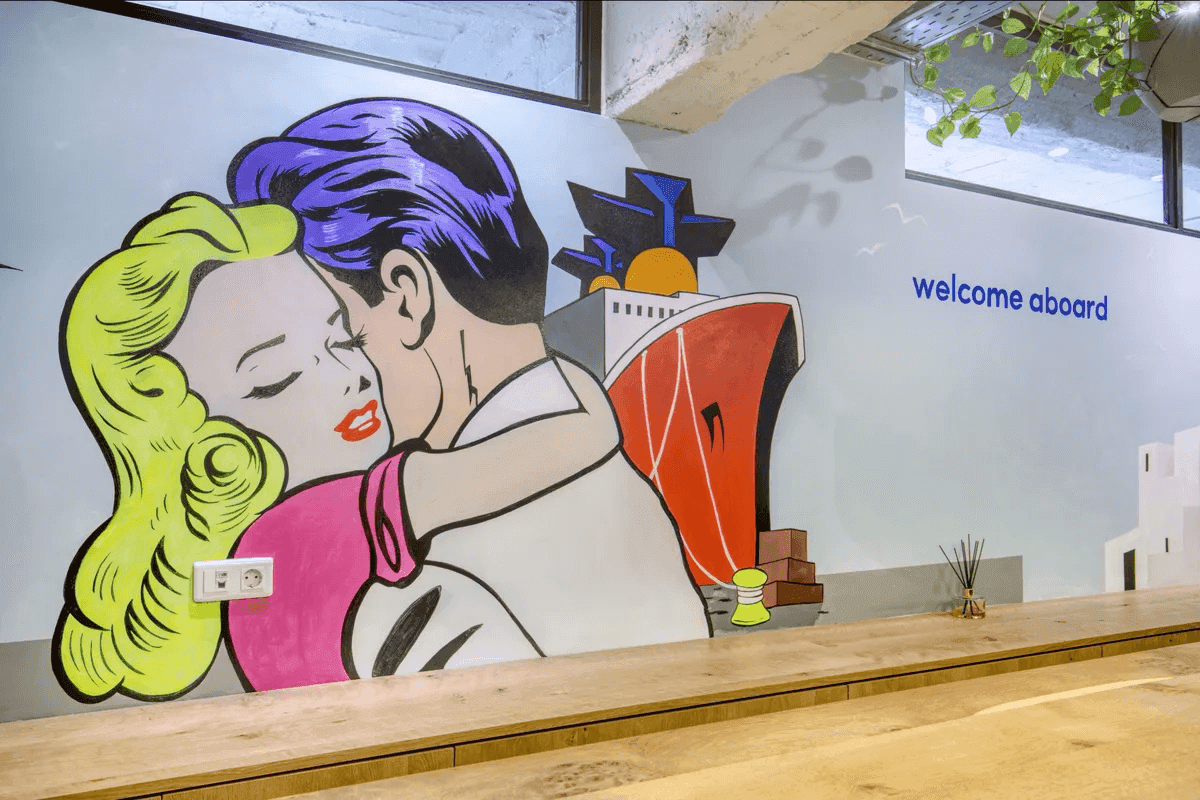 The Ferryscanner lobby where passengers can purchase tickets, featuring a sleek ticket counter and comfortable seating areas. A prominent and colorful wall art piece by artist Vasilis Michopoulos, also known as VMixo, adorns the background.