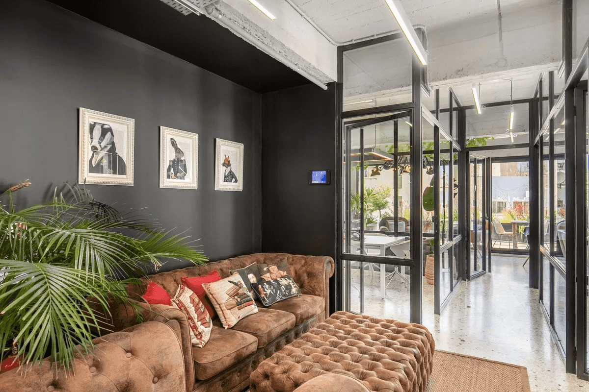 A stylish waiting area inside Ferryscanner's offices, featuring a plush brown sofa with decorative cushions, a matching ottoman, and lush green plants. The space has a modern, cozy ambiance with a black accent wall adorned with framed artwork and glass partitions leading to a bright meeting area.