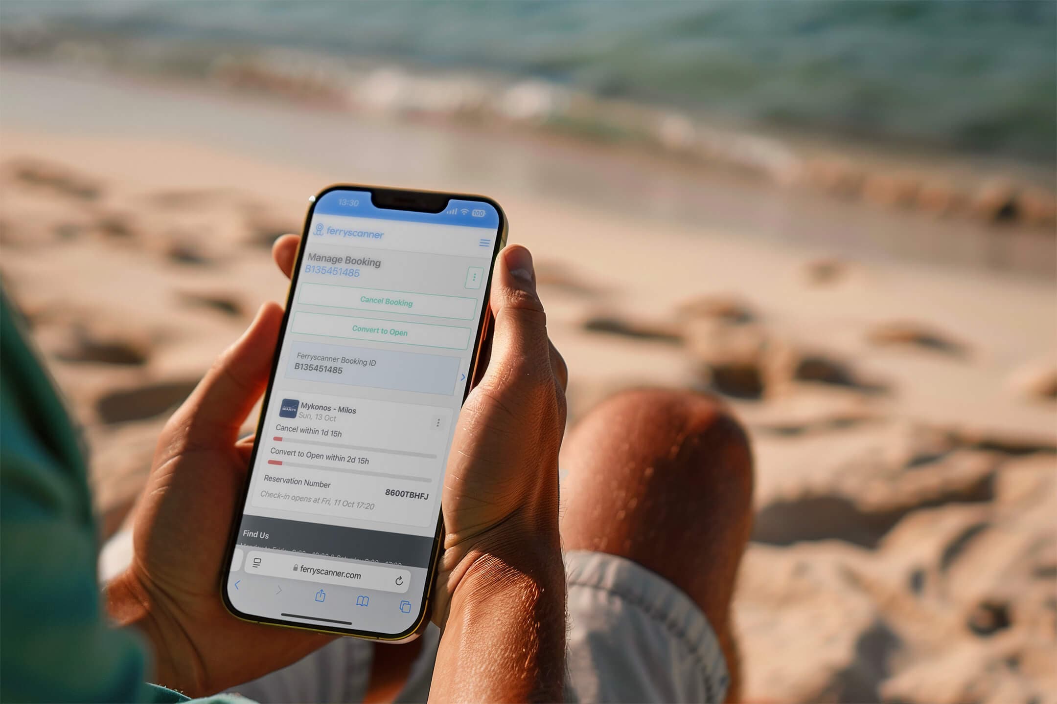 A person holding a smartphone displaying Ferryscanner's "Manage Your Booking" page while sitting on a sandy beach with the sea in the background, symbolizing convenience in managing ferry bookings anytime, anywhere.