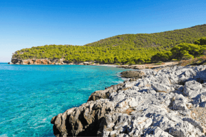 Panoramic photo of a rocky land with clear blue waters and many green trees in the background