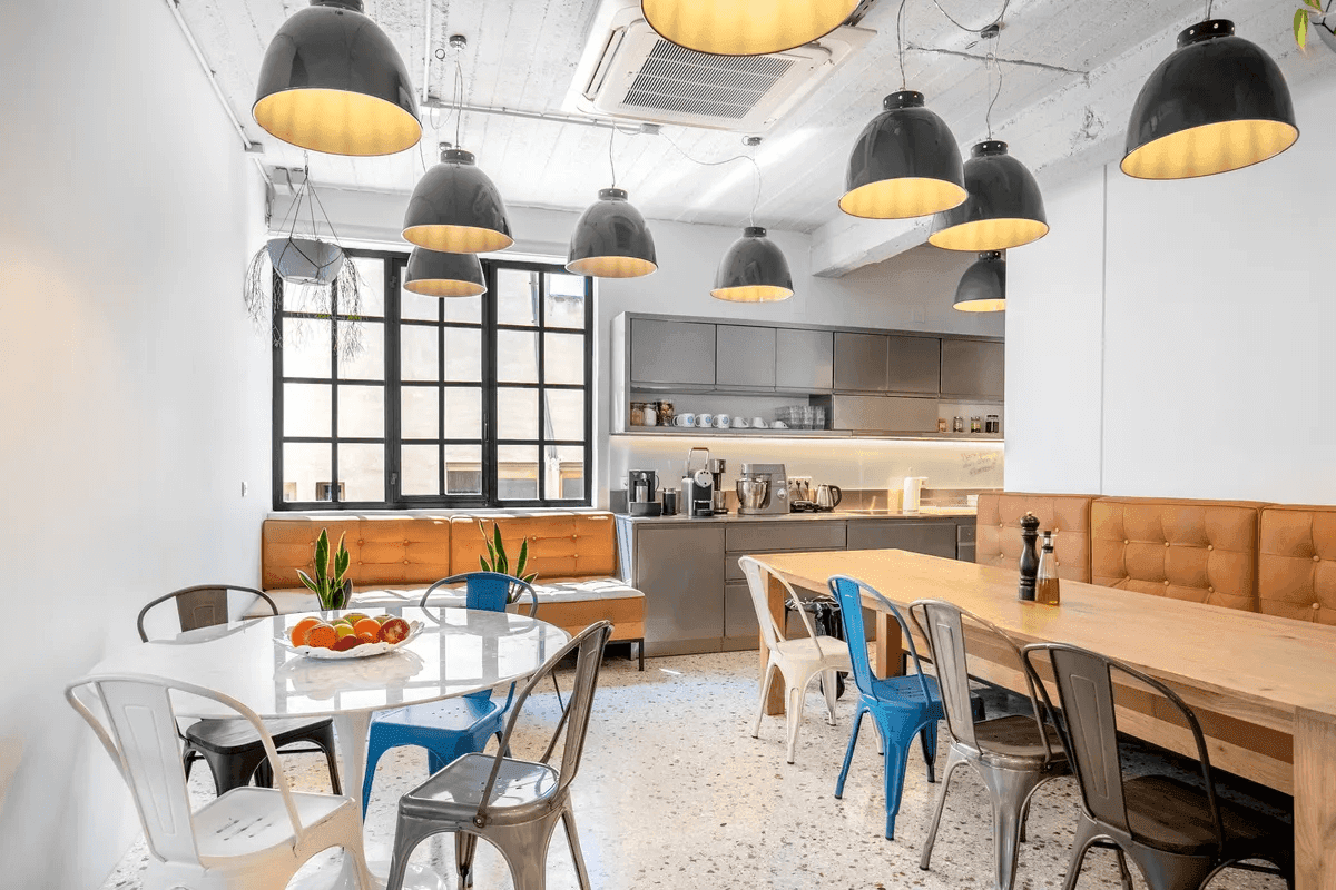 The Ferryscanner office kitchen, featuring a cozy and modern design with a mix of wooden and metal elements. The space includes a large wooden dining table with colorful chairs, a round table with metal seating, warm lighting from multiple hanging lamps, and a fully equipped kitchen area with appliances and cabinets.