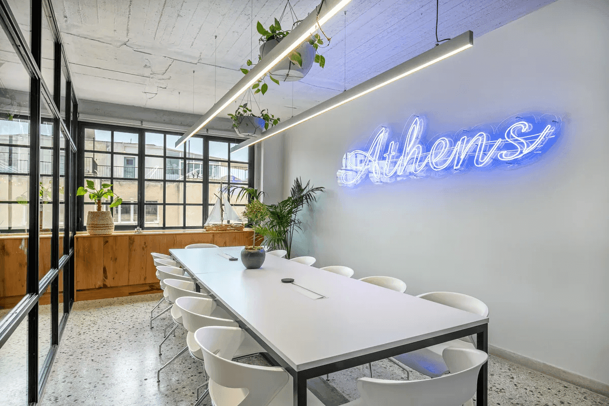 A modern meeting room in Ferryscanner's Athens office, featuring a long white conference table surrounded by white chairs, large industrial-style windows, green plants, and a blue neon sign on the wall that reads "Athens".