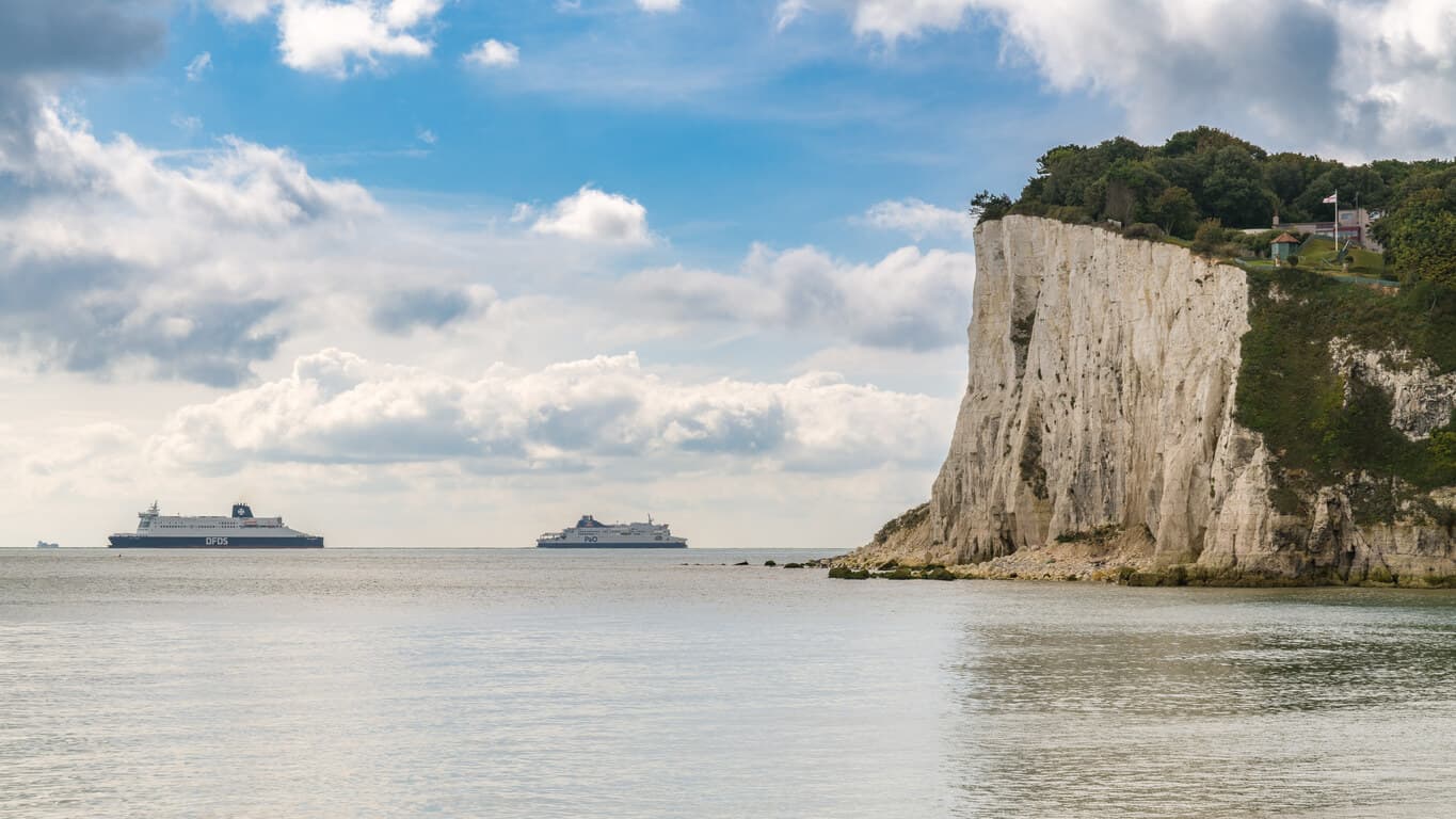 large white rock formation that is the cliffs of dover on the right side while two large ferry ships sit on the horizon sailing from france to the UK