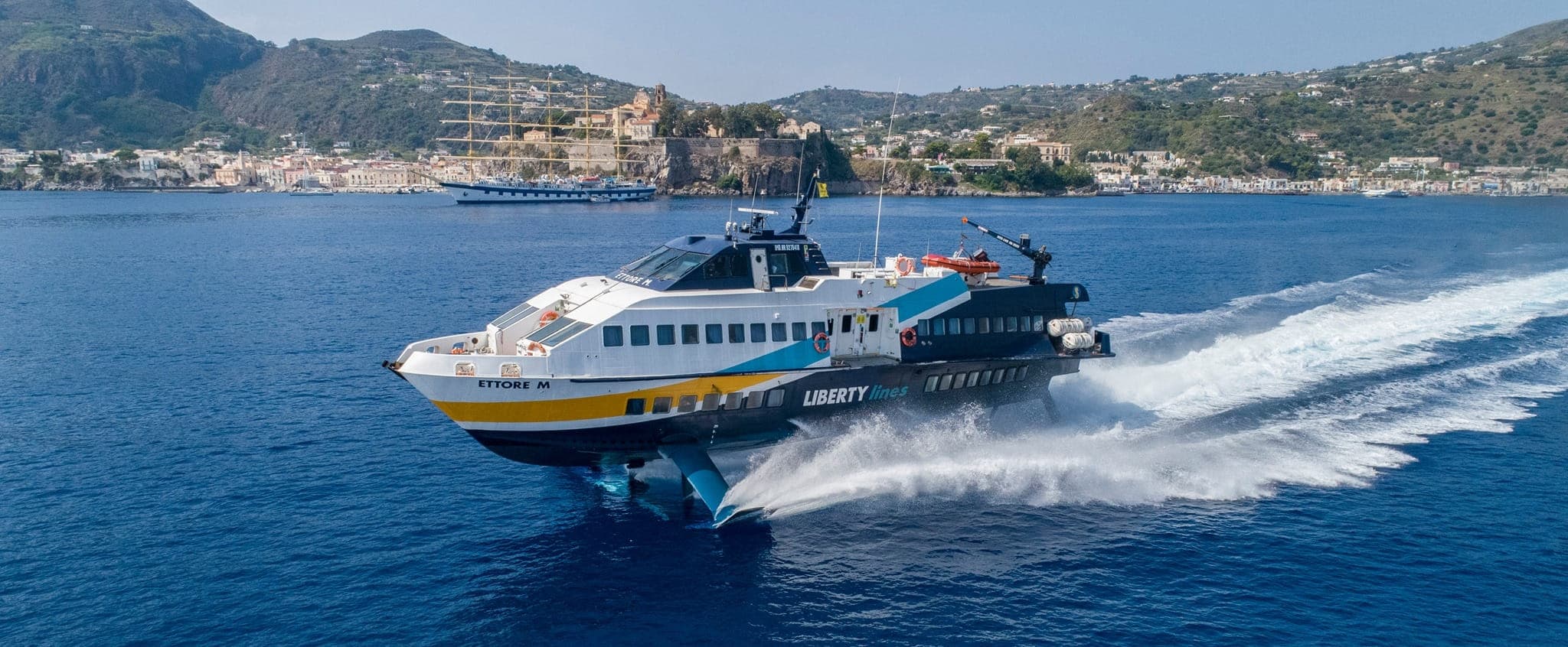 The high-speed vessel Ettore M of Liberty Lines sailing near Sicily, with a coastal town and island landscape in the background.